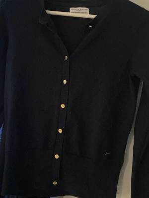 Marinblå kofta från Holly & Whyte - Stilren marinblå kofta från Holly & Whyte med guldknappar framtill. Klassisk rak modell med långa ärmar och rund halsringning. Perfekt att slänga över en t-shirt eller bära stängd för en clean look.