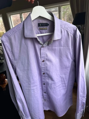 Ljuslila skjorta från Riley - Snygg ljuslila skjorta från Riley med lång ärm och klassisk krage. Skjortan har svarta och röda knappar samt en randig detalj på insidan av kragen. Perfekt för dig som vill ha en stilren look med en twist.