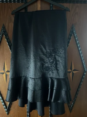 Svart långkjol med volang - Svart långkjol i glansigt material med volangkant och broderad detalj framtill. Kjolen har en dragkedja bak och en elegant, böljande form som ger en snygg siluett.