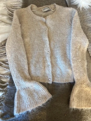 Beige fluffig kofta från AWARE - Mysig beige kofta från AWAI med fluffig känsla och rund halsringning. Koftan har pärlknappar framtill och vida, ribbstickade ärmslut som ger en unik look. Perfekt för lager på lager och riktigt skön att ha på sig under kyliga dagar.