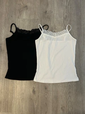 Vita och svarta linnetoppar med spets - Två stilrena linnetoppar i vitt och svart med tunna axelband och spetsdetalj upptill. Perfekta för lager på lager eller till jeans. Säljs tillsammans.