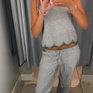 Randigt loungewear-set  - Sött randigt tvådelat set bestående av topp och byxor. Toppen har fina knytband på axlarna och en dekorativ vågig kant längst ner. Byxorna är bekväma med lös passform och dragsko i midjan. Ser ut som små blommor ut på byxorna och toppen, som insytt. Jättefint  Tröjan är i S  Byxorna är i Xs