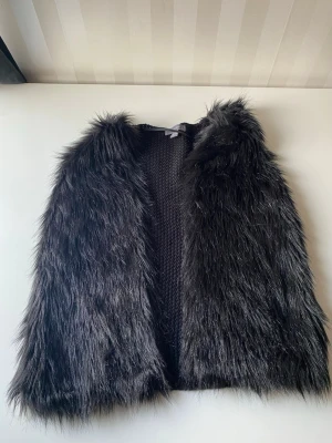 Svart pälsväst med stickad rygg från primark - Snygg svart pälsväst med lång, från primark och mjuk fuskpäls framtill och stickad baksida. Västen har en öppen design utan knäppning och är helsvart, vilket gör den enkel att matcha. Perfekt för att addera lite edge till din outfit. 