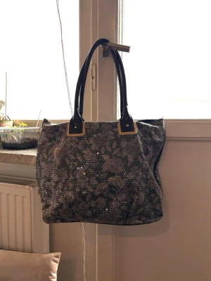 Glittrig handväska med paljetter - Snygg handväska med silverfärgade paljetter och svarta handtag. Väskan har en rymlig insida med innerfack och stängs med magnetknapp. Perfekt för dig som gillar att sticka ut. Materialet är textil täckt med paljetter och handtagen är i konstläder.