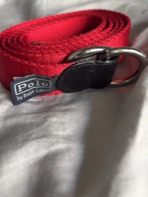 Rött bälte från Polo Ralph Lauren - Snyggt rött bälte från Polo Ralph Lauren med dubbla silverfärgade D-ringar och detaljer i svart skinn. Bältet är gjort i ett slitstarkt tygmaterial och har en klassisk, stilren design som passar till många olika outfits.