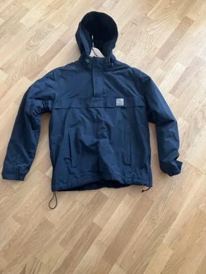 Mörkblå vindjacka från Carhartt - Fodrad på insidan,så rätt så varm.Snygg mörkblå vindjacka från Carhartt med huva och dragsko i nederkant. Jackan har en stor magficka med lock, sidofickor och en klassisk Carhartt-logga på bröstet. Tillverkad i slitstarkt syntetmaterial, perfekt för blåsiga dagar.