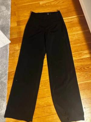 Svarta kostymbyxor från Bik Bok - Säljer ett par svarta kostymbyxor från Bik Bok i storlek 38. Byxorna har en rak och vid passform med hög midja och klassisk design. Perfekta för dig som gillar en stilren look. Materialet känns mjukt och följsamt, vilket gör dem bekväma att bära hela dagen.
