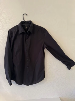 Svart slim fit skjorta från H&M - Svart skjorta från H&M i slim fit-modell med klassisk krage och knappar framtill. Skjortan är långärmad och tillverkad i ett lättskött bomullsmaterial som är easy iron. Perfekt för dig som gillar en clean och stilren look.