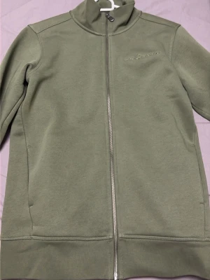 Olivgrön ziptröja från Cal Racing - Olivgrön ziptröja med hög krage från Cali racing . Tröjan har hel dragkedja framtill och diskret logga på bröstet. Materialet är mjukt och bekvämt, perfekt för en avslappnad look. Långa ärmar och ribbade detaljer vid nederkant och ärmslut.