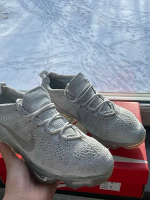 Nike Air Vapormax Flyknit vit sneakers - Nike Air Vapormax Flyknit sneakers i ljusgrått med stickad ovandel och transparent sula. Skorna har en modern design med synlig dämpning och diskret Nike-logga på sidan. Perfekta för dig som gillar en sportig och stilren look. Boxen och allt följer med.