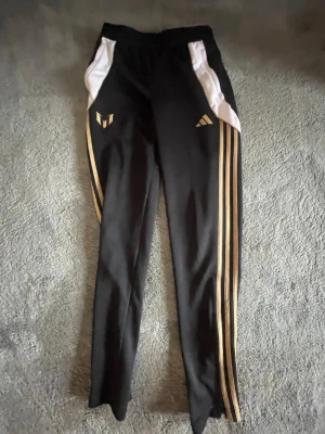 Svarta Adidas träningsbyxor Messi - Snygga svarta träningsbyxor från Adidas med guldiga detaljer och Messi-logga. Byxorna har vita partier vid fickorna och klassiska tre guldiga ränder längs benen. Perfekta för fotboll eller gymmet, med elastisk midja och smal passform.