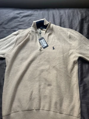 Beige halvzip-tröja från Polo Ralph Lauren - Snygg beige halvzip-tröja från Polo Ralph Lauren med klassisk blå logga broderad på bröstet. Tröjan har hög krage med dragkedja och ribbade muddar. Perfekt för en stilren och avslappnad look.