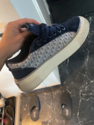 Blå Dior sneakers - Säljer ett par snygga blå Dior sneakers med klassiskt monogrammönster på sidan och vit sula. Skorna har låga skaft, mörkblå mockadetaljer och matchande blå skosnören. Finns box och kvitto