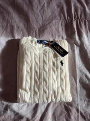 Vit kabelstickad tröja Polo Ralph Lauren - Kabelstickad vit tröja från Polo Ralph Lauren med rund halsringning och klassisk broderad logga på bröstet. Tröjan har långa ärmar och ribbade muddar vid ärmslut och nederkant. Perfekt för en clean och stilren look. Mått:  Längd: 62 cm Axelbredd: 38 cm