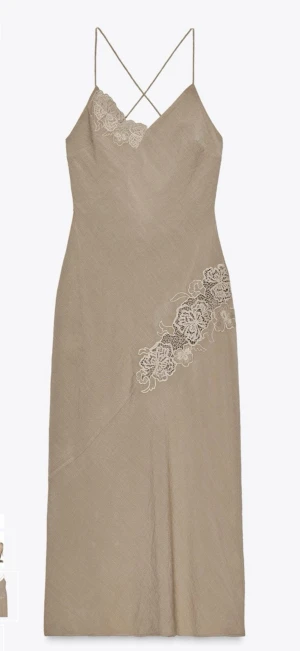 Beige klänning med spetsdetaljer - Stilren beige klänning med tunna axelband som korsas i ryggen och snygga spetsdetaljer på bröstet och sidan. Klänningen har omlott-effekt baktill med knytband och är tillverkad i ett lätt material som passar perfekt för varma dagar.