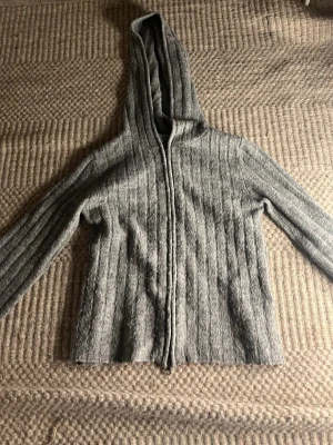 Grå stickad hoodie med dragkedja - Mysig grå stickad hoodie med dragkedja framtill och ribbade detaljer. Hoodien har långa ärmar och en stor huva som ger en avslappnad look. Perfekt för chill dagar eller när du vill ha något bekvämt och snyggt. Materialet känns mjukt och värmande.