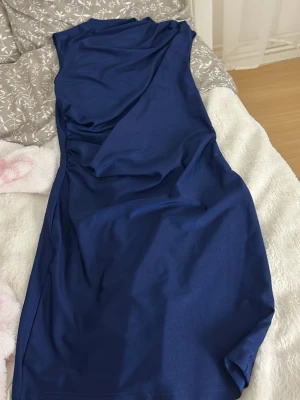 Marinblå axelbandslös klänning - Säljer en marinblå axelbandslös klänning med snygg drapering framtill. Klänningen har en elegant, figurnära passform och är tillverkad i ett glansigt polyestertyg som ger en lyxig känsla. Perfekt för dig som vill sticka ut med en stilren look.