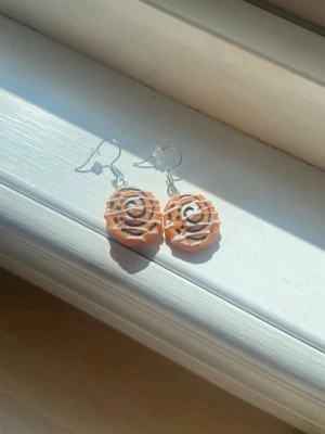 Örhängen med kanelbulle-design - Unika örhängen formade som små kanelbullar med vit glasyr. Perfekta för dig som vill ha något lekfullt och annorlunda i din stil.