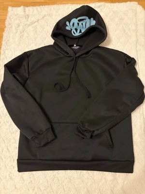 Svart hoodie med blått tryck på huvan - Snygg svart hoodie från Urban Classics med ett unikt blått graffiti-inspirerat tryck på huvan. Hoodien har dragsko i huvan, ribbade muddar vid ärmslut och nederkant samt en klassisk känguruficka framtill. Perfekt för en streetwear-look.