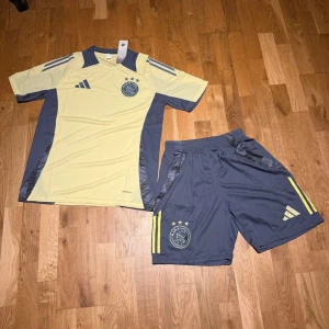 Ajax summer set ljusgul - Säljer ett Ajax fotbollskit från Adidas med en ljusgul tröja och mörkgrå detaljer samt matchande mörkgrå shorts med gula ränder. Båda delar har Ajax-loggan och Adidas-märket. Materialet är lätt och andas, perfekt för träning eller match.