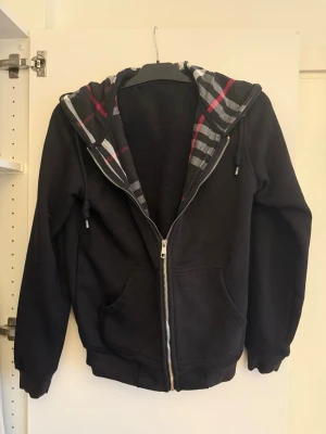 Svart burberry hoodie - Säljer en svart hoodie med dragkedja och stor huva. Insidan av huvan har ett rutigt mönster i grått, vitt och rött. Hoodien har två fickor framtill och ribbade muddar vid ärmar och nederkant. Perfekt för en avslappnad och cool stil. Pris kan diskuteras vid snabb affär