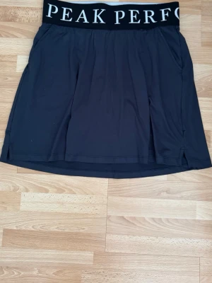 Svart skort från Peak Performance som ny - Snygg svart skort från Peak Performance med bred resår i midjan där loggan syns tydligt. Skorten har en sportig look och är gjord i ett mjukt, stretchigt material som passar perfekt för en aktiv livsstil. Hade på mig en gång.