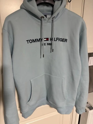 Ljusblå hoodie från Tommy Hilfiger - Ljusblå hoodie från Tommy Hilfiger med broderad logga och text 'EST. 1985' på bröstet. Klassisk känguruficka framtill och justerbar huva med dragsko. Perfekt för en avslappnad och stilren look.