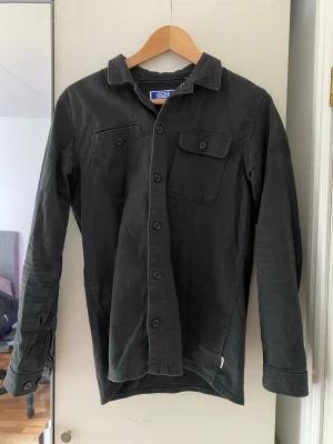 Svart overshirt från Jack & Jones - Säljer en svart skjorta från Jack & Jones med klassisk krage och knappar framtill. Skjortan har två bröstfickor med knappstängning och långa ärmar. Perfekt för dig som gillar en enkel och stilren look. Jag har tagit bort Jack & Jones loggan på armen men inget som syns tydligt . Blå med drag av grön.