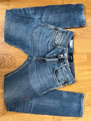 Blå jeans Replay Rocco comfort fit - Säljer ett par klassiska blå jeans från Replay, modell Rocco comfort fit. Jeansen har fem fickor, normal midja och raka ben. Materialet är slitstarkt denim i bomull med snygga slitningar och tvättade detaljer för en avslappnad look. Därimot fins det ett litet hål på knät och svaga färg stains som inte går bort men de är inget man tänker på. De passar nog inte som dom finaste byxor men skulle nog kunna nvändas som byxor man inte behöver va rädd över men ändå va feta