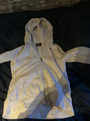Grå zip hoodie från Nike - Säljer en grå zip hoodie från Nike med klassisk huva och fickor framtill. Hoodien har dragkedja hela vägen och en liten Nike-logga broderad på bröstet. Perfekt för chill dagar eller när du vill ha en sportig look.