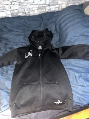 Svart hoodie från Unihoc med dragkedja - Säljer en svart hoodie från Unihoc med hel dragkedja framtill och två fickor. Hoodien har tryckta loggor på bröstet och nedtill, samt en klassisk huva. Materialet är mjukt och sportigt, perfekt för chill eller träning.