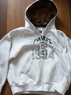 Maison Margiela x Supreme zip up hoodie - Grå zip up hoodie från Supreme och Maison Margiela. Storlek S men passar M. Den har tryck på bröstet med Maison Margiela märke och Supreme märke. Den har döskallar på insidan av huvan. Perfekt för dig som vill vara bekväm och som vill sticka ut.