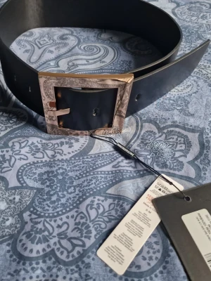 Svart bälte med stor guldfärgad spänne - Snyggt svart bälte med bred rem och ett stort fyrkantigt guldfärgat spänne. Perfekt accessoar för att lyfta din outfit med en trendig touch.