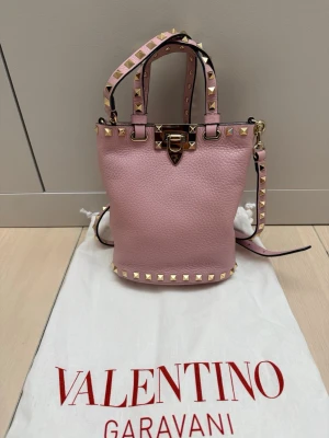 Elegant Valentino Garavani Rockstud väska i puderos  - Säljer en jättefin Valentino Garavani Rockstud-väska i en vacker, klassisk valentino-puderrosa färg med klassisk Rockstud-design. Väskan är både stilren och exklusiv – perfekt för både vardag och fest.  👜 Detaljer: - Mått: 18cm H x 17cm B x 8cm D - En sällsynt modell. Tillverkad i exklusivt  läder med klassisk Rockstud-design -Guldfärgade rockstud och detaljer - Avtagbar axelrem  -Dustbag medföljer  💗 Väskan köptes i Valentino buitken ca 1,5 år sen. Nypris: ca 15000kr. Nu: 5499kr