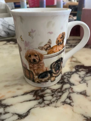 Vit mugg med hundmotiv - Söt vit porslinsmugg dekorerad med detaljerade illustrationer av olika hundvalpar och smådjur. Muggen har blommiga inslag och små bin som flyger runt motiven. Perfekt för dig som älskar hundar och vill ha en unik kopp till kaffet eller teet.