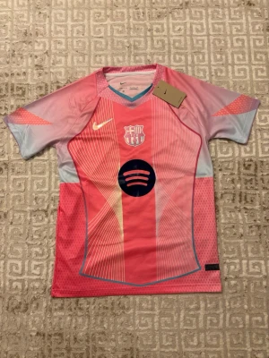 Rosa barca tröja - Säljer en FC Barcelona matchtröja från Nike i rosa och ljusblått med grafiskt mönster och klubbmärke på bröstet. Tröjan har Dri-Fit-material som andas och korta ärmar. Perfekt för dig som älskar fotboll och vill sticka ut på planen.