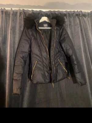 Svart pufferjacka med fuskpäls - Snygg svart pufferjacka med guldfärgade dragkedjor och fuskpäls på huvan. Jackan har två snedställda fickor framtill och quiltad design som ger en cool look. Perfekt för kalla dagar när du vill hålla stilen.