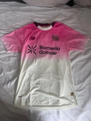 Bayer Leverkusen rosa matchtröja - Snygg Bayer Leverkusen matchtröja från New Balance i rosa och vitt med gradient-effekt. Tröjan har klubbmärke och sponsortryck på bröstet samt rund halsringning med mörklila kant. Tillverkad i lätt och ventilerande funktionsmaterial.