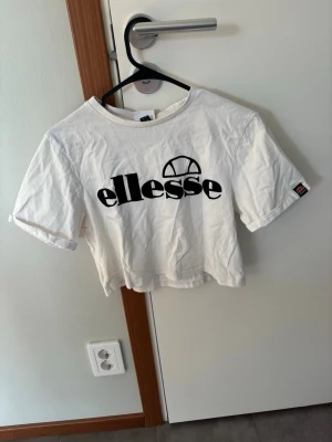 Vit croppad t-shirt från Ellesse - Snygg vit croppad t-shirt från Ellesse med svart logga framtill. T-shirten har korta ärmar och en relaxed passform. Perfekt till jeans eller shorts för en chill och sportig look. I storlek 36🤍