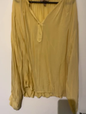 Gul blus med knappar från H&M - Ljusgul blus från H&M med v-ringning och dekorativa knappar framtill. Blusen har långa, lösa ärmar och är tillverkad i ett tunt, flowy material som ger en avslappnad look. Perfekt att styla med jeans eller kjol.