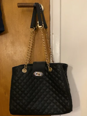 Svart quiltad handväska med guldkedjor - Stilren svart handväska i quiltat konstläder med guldfärgade kedjehandtag och vridlås. Väskan har rymligt innerfack med dragkedja och flera mindre fack för organisering. Perfekt för dig som gillar klassisk och trendig design.