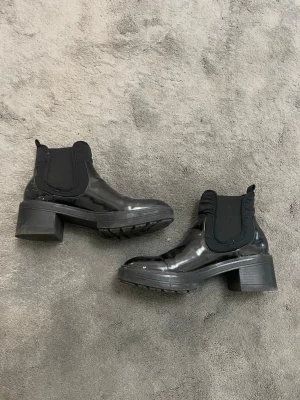 Svarta lackade Chelsea boots från Zara - Säljer ett par svarta Chelsea boots från Zara i glansigt lackmaterial. Skorna har bred resår på sidorna, rundad tå och kraftig klack. Perfekta för dig som vill ha en edgy och trendig look till hösten.