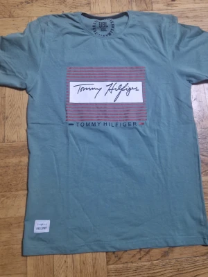 Grön Tommy Hilfiger t-shirt XXL - Snygg grön t-shirt från Tommy Hilfiger i storlek XXL. Framsidan har ett tryck med röda ränder och logga i vitt och svart. Rund halsringning och korta ärmar. Tillverkad i mjuk bomull för skön känsla. Liten patch med 'INDO SPIRIT' nere till vänster.