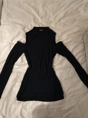 Svart långärmad topp med cut out - Trendig svart långärmad topp med cut out-detaljer vid axlarna och hög hals. Tight passform som framhäver figuren. Perfekt för dig som vill ha en edgy och stilren look. Materialet känns mjukt och stretchigt, troligen polyester eller bomullsmix.