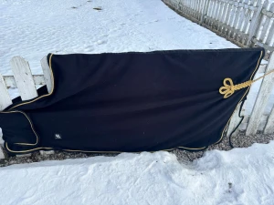 Svart fleecetäcke med guldkant - Säljer ett svart fleecetäcke från Back on Track med snygg guldkant och dekorativt snöre. Täcket har kardborreknäppning och är perfekt för häst. Materialet är mjukt fleece och täcket har en klassisk design.