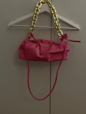 Rosa handväska med guldkedja - Rosa handväska i mjukt material med snyggt rynkad design. Väskan har en rejäl guldfärgad kedja som handtag och en lång axelrem i matchande färg. 