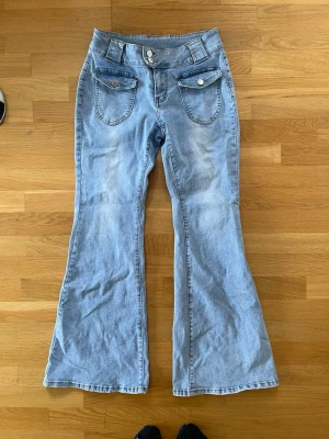 Ljusblå bootcut jeans med fickdetaljer i storlek M - Säljer ett par ljusblå bootcut jeans med dubbla knappar i midjan och stora fickor framtill med vita knappar. Jeansen har utsvängda ben och klassisk femficksdesign baktill. Storlek medium. Perfekta för en retroinspirerad look!