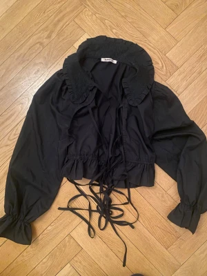Svart blus med volangkrage och knytband - Trendig svart blus från Glamo med volangkrage och långa knytband framtill. Blusen har snöre i midjan och vid ärmsluten för en puffig effekt. Tillverkad i ett lätt tyg som ger en luftig känsla. Perfekt för dig som gillar statement-detaljer. Storlek 40 men skulle säga att den passar en 36/38 för en lite oversize-känsla. 