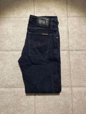 Mörkblå jeans från Nudie Jeans - Säljer ett par mörkblå jeans från Nudie Jeans med klassisk femficksmodell och snygga sömdetaljer på bakfickan. Jeansen har normal midja och raka ben, tillverkade i slitstark denim. Perfekta för dig som gillar stilrena och tidlösa jeans.