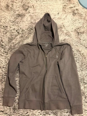 Mörkgrå hoodie från Polo Ralph Lauren - Snygg mörkgrå hoodie från Polo Ralph Lauren med dragkedja framtill, två fickor och klassisk huva med dragsko. Tillverkad i mjuk bomullsblandning och har den ikoniska lilla loggan broderad på bröstet. Perfekt för en chill och stilren look.